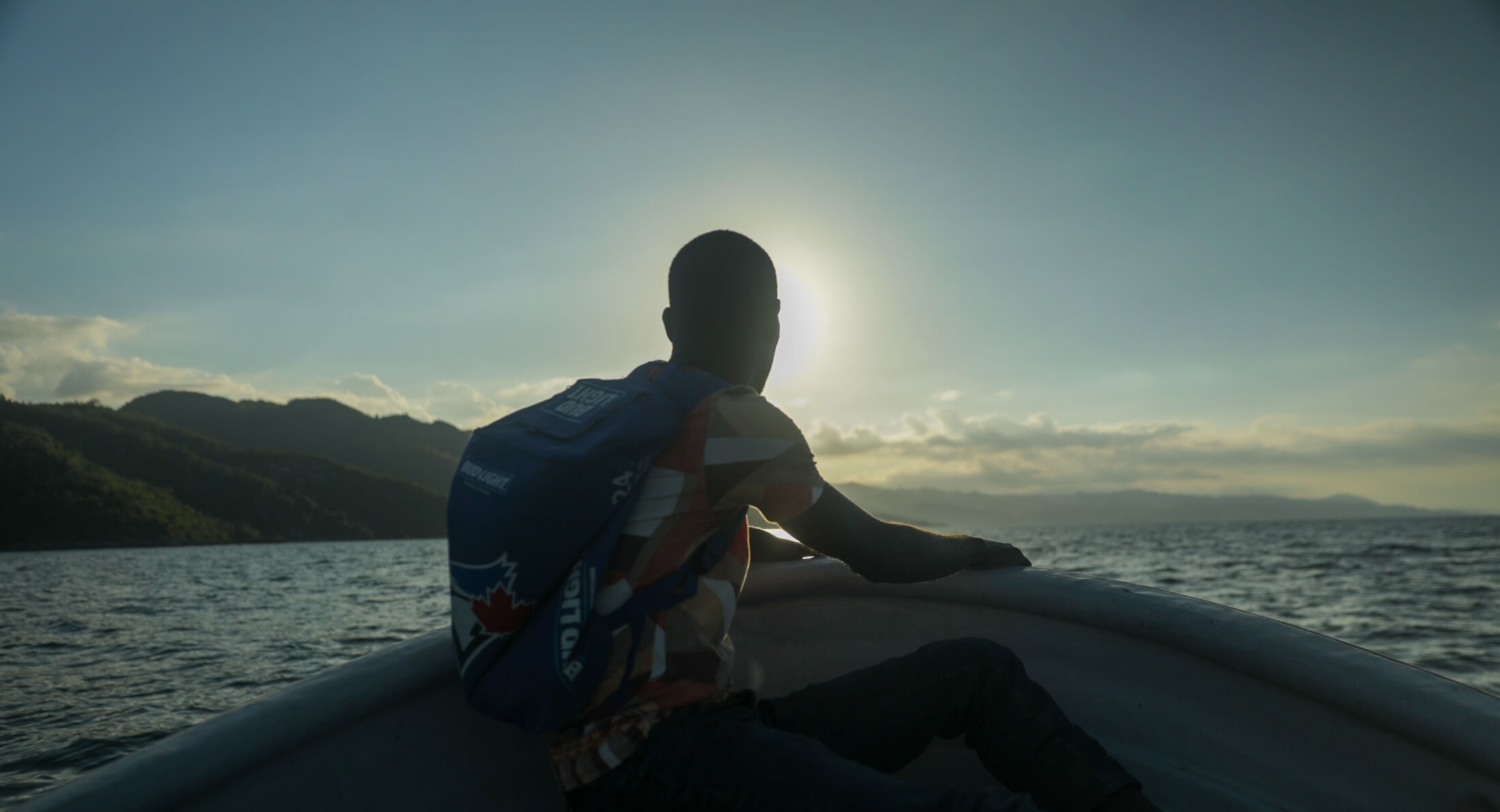 un homme assis à l'avant d'un petit bateau, avec l'océan et des montagnes en arrière-plan